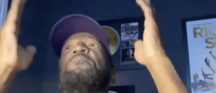 Man gesturing with hands raised, wearing cap.